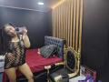 EmilyVidal in a black fishnet dress, showing her tits, holding a phone, standing in a bedroom with a gold-trimmed black bed, red blanket, and wooden wall.