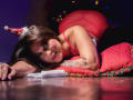 LindaJimenex, wearing a red Santa hat and sheer red dress, is on all fours with a tattooed arm, smiling at the camera, her red-assed butt raised.