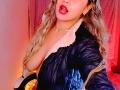 AnastaciaMonaco with wavy blonde hair, red lipstick, black puffy jacket with white fur, and visible large tits, tongue slightly out, in a pink-lit room.