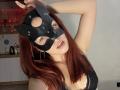 AlexXas with red hair, wearing a black leather mask with ear points, hoop earrings, and a black lace bra, posing with arms up, showing her tits and open mouth.