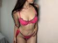 Tisia with long dark hair, pink lace bra with floral trim, and pink lace panties, standing, showing her tits and pussy, hands near her hips, light skin, leaning slightly.
