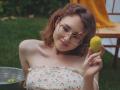 TeresaBirt with curly brown hair, glasses, and a floral off-shoulder top, holding a yellow lemon, smiling, showing her small tits, standing on green grass, and metal bucket.