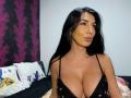 BrunetteBabe69 with big tits in a black lace bra, pouty lips, long black hair, and tan skin, sitting on a bed with colorful pillows.