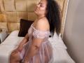 KloheEvans with curly hair, wearing a sheer pink dress, sitting on a bed, showing her ass and tits, looking up with a slight smile, in a bedroom.