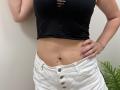Tilliana has a black, cut-out, sleeveless top showing her tits and a flat, tan belly. She's wearing white, button-up pants with pink, glittery nails and a ring.