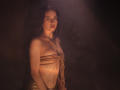 TriciaBarnes with bare tits, bound in rope, standing in dim light. She has long brown hair, a tattoo under her tits, and a small belly button piercing.
