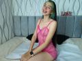 AbbyHillsI with green and black hair, smiling, wearing a pink sheer lingerie dress, revealing small tits, sitting on a bed, wearing a smartwatch, looking cute.