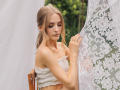 EvaDaly with small tits, wearing a white, off-shoulder top, gently holding a lace curtain, blonde hair in a ponytail, looking down, in a soft outdoor setting.