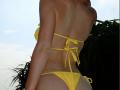 PeacHub69's back, showing a yellow bikini with strings, sand on her tan skin, and her ass, standing with green palm leaves in the background.