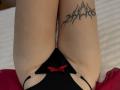 FallenAngelia's pale thighs, black thong with red bow, and black tribal tattoo on right thigh, sitting with knees together. Visible black straps on sides. Red fabric below.