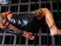 NenaBlack is lying on a metal grid with her ass up, wearing a black leather dress, red bra, and tattooed arms and legs. Her wrists are cuffed.