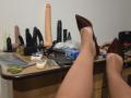 BeccaVanBoa's legs with brown pointed heels on a desk, surrounded by dildo toys, keys, scissors, phone, and a box of "KILLS" in a cluttered room.