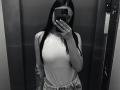 Black-and-white mirror selfie of Olieri with long dark hair, wearing a white sleeveless top, showing small tits, tattoo on right arm, and jeans, holding phone, covering face.