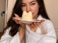 EmeraldCrivaro with long brown hair, wearing a white fluffy robe, holding a cat-shaped cream-colored ceramic thing in her mouth, looking at the camera.