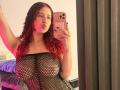 ValeryScot, con i capelli rossi e ricci, labbra piene, indossa un abito nero in rete, che mette in mostra il suo seno generoso, tiene in mano un telefono rosa, in un selfie allo specchio..