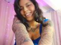 Chili, smiling, has dark wavy hair, blue tank top, and tattoos covering her arms. She's showing her tits and has a necklace. Pink lights are in the background.