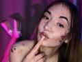 LelaTraverso, with long brown hair and tattoos, pokes her pink-lipsticked, slightly glistening, bare-chested face with her finger, under purple and pink lighting.