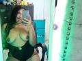 AriaVega with curly black hair, sticking out tongue, topless with large, natural tits, wearing black high-waisted bottoms, taking mirror selfie, holding phone, green mirror reflection.