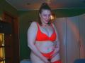 VeronikaGoldy, plus-sized with pale skin, red bra and panties, standing, hand on her fat pussy, smiling, brown hair in a ponytail, green walls, white closet, blue bed.