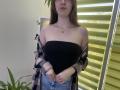 Nesie with long brown hair, wearing a black strapless top, pink and black plaid shirt off her shoulders, and blue jeans, standing in front of yellow blinds.