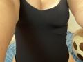 Olieri's black tank top is tight, showing her boobs and cleavage. She's wearing it with black panties, and her tan arms are visible. Soft blue and brown background.
