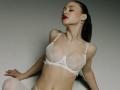 MilagrosEdmisten with her eyes closed, wearing a white lace bra and matching panties, has red lipstick, a navel piercing, and her dark hair is pulled back. Boobs are visible.