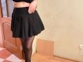 EmmaNoa indossa una gonna nera, collant neri trasparenti e un top corto nero, con un tatuaggio sul lato destro, in piedi su un pavimento di legno, mostrando il suo sedere..