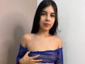 NaomyRosse with long black hair, wearing a sheer blue off-shoulder top, covering her small tits with one hand, looking at the camera with red lipstick.