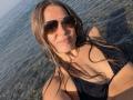 AuroraStar with wet brown hair, wearing aviator sunglasses and a black bikini top, smiling at the camera in shallow water. Her boobs are visible and she's looking sexy.