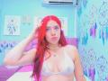MiaLeroy with bright red hair, wearing a sheer white bra, showing her tits, a shell necklace, and a flower in her hair, in a colorful room.