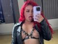Estefy69 with red curly hair, black leather jacket, and black strappy harness, sticking her tongue out, holding a purple phone, showing her tits with chains.