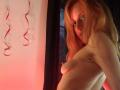 Topless LyannaDifran with pale skin and red hair, small tits, and a side glance, looking slightly annoyed. She's in dim red lighting with black curtains in the background.