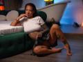 RosaInk with brown skin and tattoos, wearing a white crop top and plaid skirt, sitting on the floor, legs spread, finger in mouth, green bed behind her.