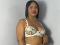 AliceBooty with medium brown skin, black bob, red lipstick, wearing white lace bra with floral embroidery, showing her tits, standing against white wall, smiling, wearing gold necklace.