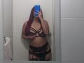 AntoniaBrunni with red hair, covering her face with a blue phone, wearing black strappy lingerie, showing her tits and glistening pussy, standing in a bathroom mirror selfie.