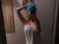 Olieri with blue towel on head, white towel covering her tits, holding phone, mirror selfie, tattoo on right shoulder, pink nails, wood door on left, dim lighting.