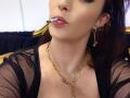 VanessaSquirtt with a cigarette in her pouty lips, wearing a sheer black top, gold chain necklace, and dangling earrings, has her tits slightly visible and her ass is off-screen.