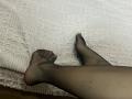 Les jambes de Destinis, habillées de collants noirs transparents, sont légèrement croisées sur un tapis blanc texturé. On peut voir ses orteils, et il y a une petite cicatrice sur son mollet droit..