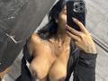 MajoJones with wet tits, large dark nipples, wearing a black jacket, taking a mirror selfie, holding a phone with sparkles, showing tattoos on her arm and chest.