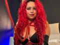 Estefy69 with bright red curly hair, black bra, red leather harness, and black gloves, holding a red rope, smiling, showing her large boobs.
