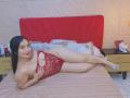 MelaniCoox with red lace lingerie, showing her big ass and small tits, lying on a bed, legs crossed, smiling, black hair, tan skin, wearing white sandals.