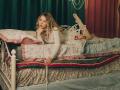 KylaHemeke with wavy blonde hair, laying on a white metal bed, wearing a white long-sleeve dress, holding a bead necklace, legs up, looking at camera.