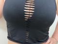 Tilliana's black, cut-out, sleeveless top with a vertical row of oval holes exposing her tan, cleavage-filled, large, round, bare breasts. Her right hand rests on her hip.