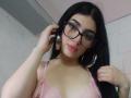 IsabellaaRossi with black hair, glasses, pink lace bra, and large hoop earrings, holding phone to ear, looking down with pink lipstick. Boobs visible through lace.