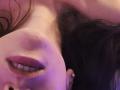 MiaReed's face, close-up, showing pale skin, dark brown hair, purple-lit lips, slight smile, small gold earring, and visible teeth, lying down with head tilted.