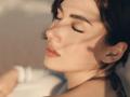 OliviaCluff's face is in soft sunlight, eyes closed, with her nose and lips highlighted. She has smooth skin, brown hair, and subtle makeup. Her right arm is slightly visible.