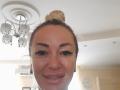 MysteryLaddy has blonde hair in a bun, thick eyebrows, long eyelashes, and is smiling with slightly flushed cheeks. A chandelier is above her head.