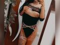 DenyChantal with long black hair, wearing black lace top, exposing her tits, black and pink garter lingerie, tattooed thigh, holding phone in mirror selfie.