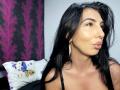 BrunetteBabe69 with pouty lips, black hair, and shiny skin, wearing a black strap top and long earrings, looking to the side. Boobs visible, pillow with blue bird in background.