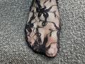 Tattiya's foot in black lace with floral patterns, showing her toes through the semi-transparent fabric, resting on a textured gray carpet.